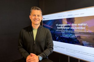 Mann med kort, mørkt hår smiler mot kamera. Han har på seg svart genser med en lys t-skjorte synlig i åpningen. En TV står på i bakgrunnen, inne på kontoret hans.