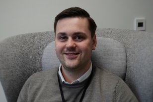 Mann i grå genser som sitter i en grå stol med et svart arbeidskort rundt halsen og smiler til kamera.