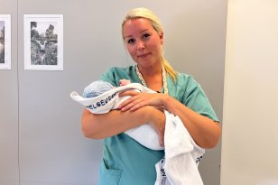 Kvinne med langt blondt hår samlet i strikk bak, iført turkis/grønn arbeidsuniform, holder en nyfødt baby pakket i hvit pledd foran kamera.
