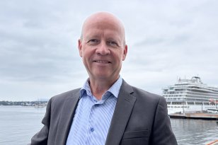 Mann i mørk grå dressjakke og blå skjorte står foran kamera. I bakgrunnen ses et cruiseskip og havet under overskyet himmel.