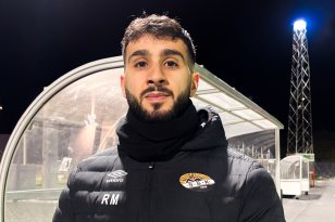 Mann med svart hår, skjegg og bart poserer foran en fotballbås utendørs om kvelden. Han har på seg en langermet jakke, og lys fra stolper skimtes i bakgrunnen.