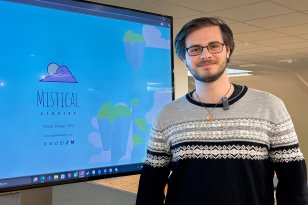 Mann med kort, mørkt hår og briller står foran en TV-skjerm på et kontor. Han har på seg en langermet genser i svart og grått. I bakgrunnen skimtes et felles kontorområde.