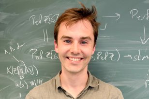 Portrett av Matematiker. En mann med brunt, kort hår og grønn, kortermet t-skjorte står foran en grønn tavle. Bak ham er det skrevet matematiske formler. Han ser mot kamera og smiler.