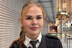 Kvinne med langt oransjegult hår i strikk smiler til kamera. Hun har på seg hvit skjorte, slips og mørkegrå genser som del av vekteruniform. I bakgrunnen skimtes deler av arbeidsplassen på Gardermoen.