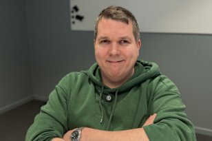 En mann sitter ved et bord med armene krysset foran seg og ser mot kamera med et svakt smil. Han har kort, gråsprengt hår og er kledd i en grønn hettegenser. På håndleddet har han en klokke med metallrem. Bak ham er en grå vegg med et enkelt kunstverk i hvitt og svart.