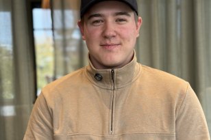 En ung mann med svart caps med Pointer-logo og beige genser med glidelås i halsen smiler mot kameraet. I bakgrunnen er det store vinduer med grønne gardiner.