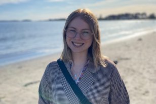 En kvinne smiler mot kameraet på en solfylt strand. Hun har briller, skulderlangt blondt hår og rutete frakk. Bak henne skimtes sjøen, himmelen og solskinnet.