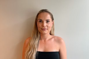 En dame med langt, blondt hår står inntil en hvit vegg og smiler med munnen lukket. Hun har på seg en svart tubetopp og et beige skjørt.