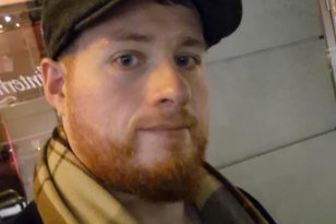 En mann med rødt skjegg tar et nærbilde av seg selv utendørs i mørket. Han har på seg en mørk caps, et rutete skjerf og en mørk jakke. I bakgrunnen er det en bygning med opplyste vinduer.