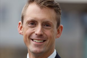 En mann i uniform står ute og smiler varmt mot kamera. Han har kort, blondt hår gredd til siden og lyseblå øyne. Han bærer en mørk marineblå kabinpersonale-uniform med hvit skjorte og stripete slips i rødt, hvitt og blått. På jakkeslaget sitter et lite rødt merke og et vingemerke med Norwegian-logoen.