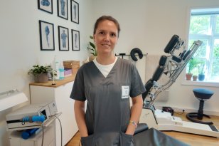 En behandler i grå klinikkuniform står i et lyst behandlingsrom med medisinsk utstyr, behandlingsbenk og anatomiske plakater på veggen. Hun smiler mot kamera.