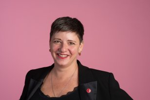 En kvinne med kort, mørkt hår smiler mot kamera. Hun har på seg en svart blazer med en rød knapp på jakkeslaget, gulløredobber og en sølvkjede. Bakgrunnen er ensfarget rosa.