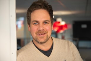 Et nærportrett av en mann som lener seg lett mot en hvit vegg. Han smiler svakt, og bakgrunnen består av et kontormiljø med røde lamper som gir varme farger.