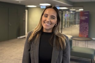 En dame med langt brunt hår. Damen har på seg en svart genser og en grå blazer. Hun står i et åpent rom med grønne vegger. Hun smiler med tennene. 