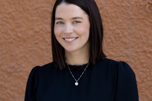 Ei dame med mørkt hår. Hun har svart kjole og et sølvsmykke på seg. Hun ser rett i kameraet og smiler. Hun står foran en brun vegg.
