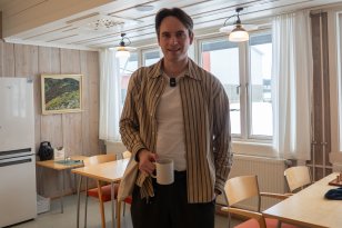 En mann står i et personalrom med lys trepanel og store vinduer. Han holder en kaffekopp i høyre hånd og ser mot kamera. Rundt ham står bord og stoler, og rommet har en rolig og uformell atmosfære.