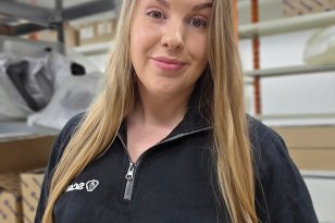 En dame med langt blondt hår smiler, hun har på seg en svart genser med glidelås som er litt åpen. Bak henne er det et stativ med ulike esker.