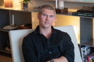 En mann med kort blondt hår sitter i en hvit lenestol og kikker mot kameraet. Han har på en svart skjorte og sitter med armene i kors. Bak han står det en hylle som er fylt med lys, blomster, lamper og bøker. 