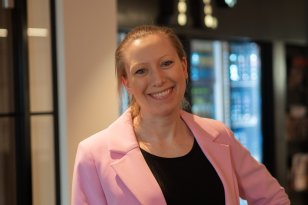 En kvinne smiler til kamera. Hun har på en rosa blazer, og står i et rom med et stort kjøleskap med glassdører i bakgrunnen.