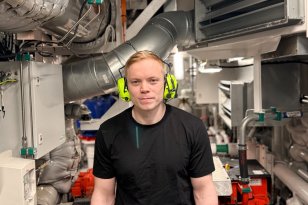 En mann står i et trangt maskinrom omgitt av store motorer, rør, ventilasjonssystemer og teknisk utstyr. Han har på seg hørselsvern og arbeidsklær, og ser rett mot kameraet. Rommet er tettpakket med maskineri og metallrør i ulike størrelser.