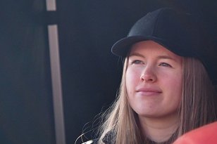 En ung dame med brunt hår og svart caps står foran en mørk vegg