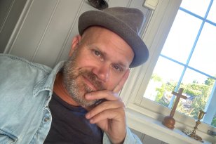 Portrett av en mann med hatt og skjegg i svart t-skjorte og dongerijakke