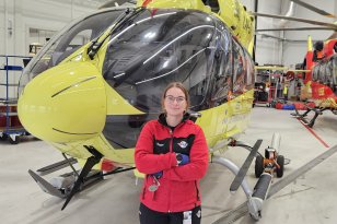 En kvinne med briller og rød arbeidsjakke står med armene i kors foran et stort, gult helikopter i en lys hangar. Et mindre rødt og gult helikopter og et svart og rødt kjøretøy står lenger bak.