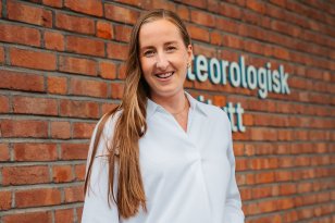 Meteorolog Pernille Borander En kvinne med brunt hår, hvit skjorte og olabukser smiler til kamera. Hun står foran en rødbrun teglstegnsvegg med logoen og navnet til Meteorologisk institutt i bakgrunnen.