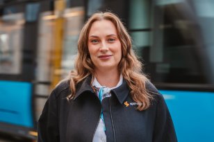 Trikkefører Ingrid Nansve En kvinne i står foran en trikk i bevegelse og smiler til kamera. Hun har på seg trikkefører-uniform med nøkkelkort rundt halsen, har brunt hår og ring i nesa.