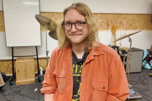 En person sitter i et musikkrom og ser inn i kameraet. Han har skulderlangt blondt hår, briller og har på seg en åpen skjorte med en band-t-skjorte under. Bak han står et trommesett, en forsterker, gitarer og andre instrumenter. Veggene er dekket av sponplater, og et whiteboard står til venstre i bildet.