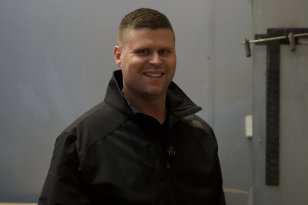 En mann smiler mot kameraet. Han står i et rom med stålvegger og har på seg en svart jakke med høy krage. På veggen til høyre henger en stålkonstruksjon