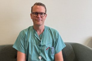 En mann i grønne sykehusklær sitter på en sofa og ser mot kamera. Han har korte, lyse hår, briller og ID-kort rundt halsen. Bak ham er en lys vegg med et innrammet bilde.