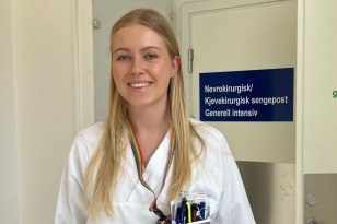 En ung kvinne i hvite sykehusklær står i en korridor på sykehuset og smiler mot kamera. Hun har langt, blondt hår og navneskilt festet på brystet. Bak henne er en dør med skilt der det står «Nevrokirurgisk/Kjevekirurgisk sengepost Generell intensiv».