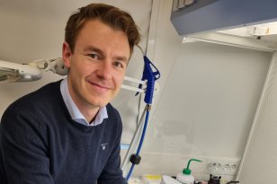 En mann sitter ved en labbenk med blå engangshansker og holder et sølvfarget optisk instrument. Rundt ham ligger presisjonsdeler og laboratorieutstyr, og han smiler mot kameraet.