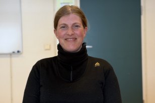 En kvinne smiler mot kamera. Hun har på seg en svart genser med høy hals og har håret trukket bakover. I bakgrunnen ser man et enkelt rom med en grønn dør og en tavle på veggen.