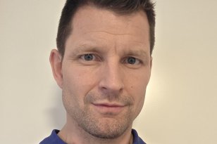 En mannlig behandler i blå poloskjorte står foran en lys, ensfarget vegg. Han ser rett mot kamera med et rolig og profesjonelt uttrykk. Han har kort, mørkt hår og lett skjeggstubb.