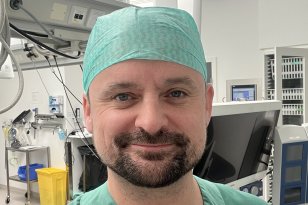 Kjetil smiler til kameraet iført grønt operasjonstøy og hette. Bak ham skimtes medisinsk utstyr og monitorer i et rent og lyst operasjonsrom på St. Olavs Hospital.