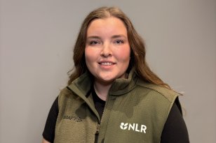 Dame med brunt hår og grønn vest over en svart t-skjorte smiler til kamera