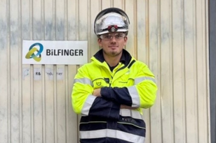 Plastmekaniker Simen Lauritzen En mann står i gul og svart arbeidsdress og lener seg mot en industrivegg. Han har på seg vernebriller, og på hodet har han en hjelm. Til venstre for han er et skilt der det står "Bilfinger".