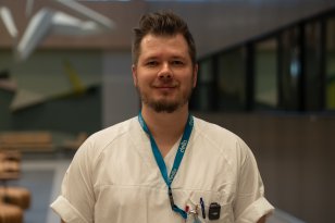 En mann står innendørs i et større bygg med åpent rom og belysning i taket. Han har på seg hvite arbeidsklær og et nøkkelkort i snor rundt halsen. Bak ham er det gangarealer og åpne soner.