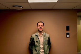 En mann i grønn jakke står inntil en oransje vegg. Han smiler og ser inn i kamera.