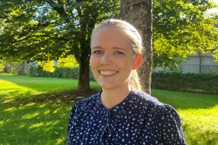 En kvinne i blå skjorte og lyst hår i hestehale står i en park med grønn plen og store trær