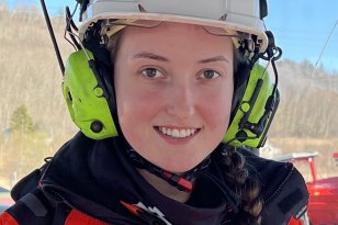 En jente smiler, har på seg hvit vernehjelm og grønne hørselsvern. Hun har en oransje jakke. I bakgrunnen sees et skogholdt og blå himmel. 