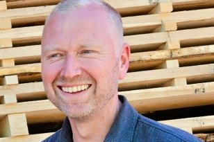 En smilende mann står utendørs foran stablede treplanker. Han har kort, lyst hår, skjeggstubb og mørk skjorte, og ser litt til siden med avslappet uttrykk.