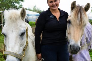 Ei dame i mørke klær. Hun står mellom to små hester. Hun er ute. 