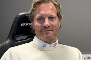 En mann med lyst, kort, bølgete hår og skjegg sitter i en svart kontorstol og ser mot kameraet. Han har på seg en lys skjorte under en hvit genser.
