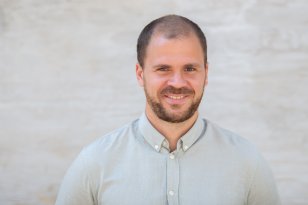 En mann i midten av 30-årene står foran en lys, nøytral bakgrunn og ser rett inn i kamera. Han har kort mørkt hår, kort skjegg og et vennlig smil. Han har på seg en lys, knappet skjorte med krage. Bildet er et klassisk portrettbilde med rolig bakgrunn og naturlig lys.