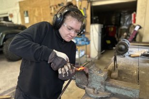 En person arbeider med et oppvarmet metallstykke som er festet i en skrustikke på en arbeidsbenk. Personen bruker verneutstyr som hansker, hørselsvern og beskyttelsesbriller i et verkstedmiljø.
