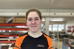 Dame med svart og oransje t-skjorte. Hun ser rett i kamera og smiler. Hun er i et verksted.