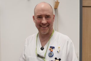 Skallet mann med hvit sykepleier uniform. I uniformslommen er det ulike figurer. Mannen har hendene i lommene på uniformen. 